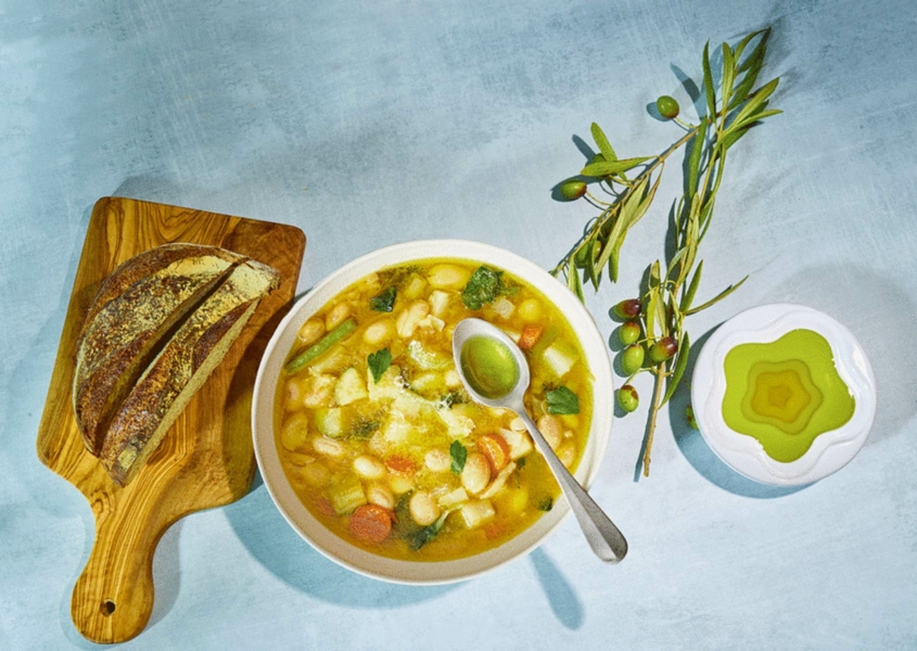 Bowl of bean suup with plate of olive oil, olive sprigs, and bread on cutting board