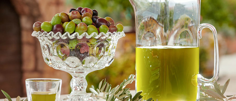 Olive oil in pitcher and glass next to glass bowl of olives